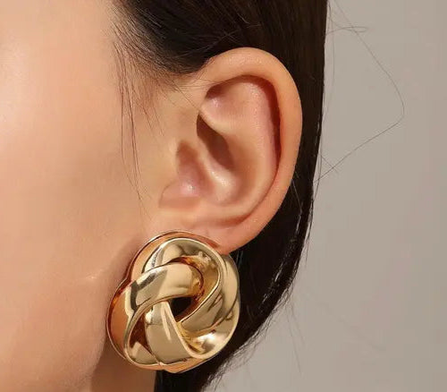 Close-up of a woman's ear wearing a large, round, gold-tone knot stud earring.