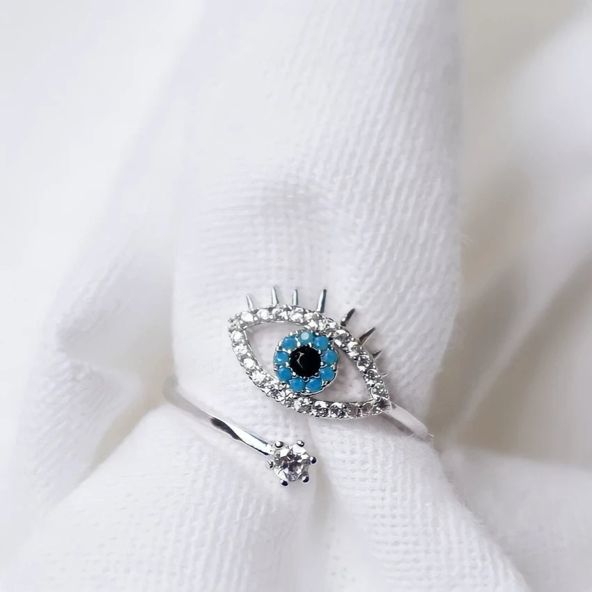 Silver evil eye ring with turquoise and clear stone detail on a white textured background.