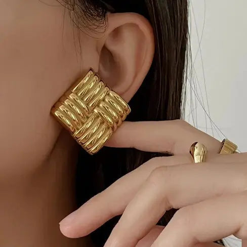 Close-up of gold woven square earrings and gold rings worn by a model.