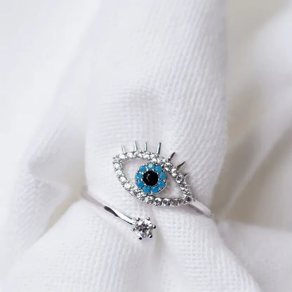 Silver evil eye ring with turquoise and clear stone detail on a white textured background.